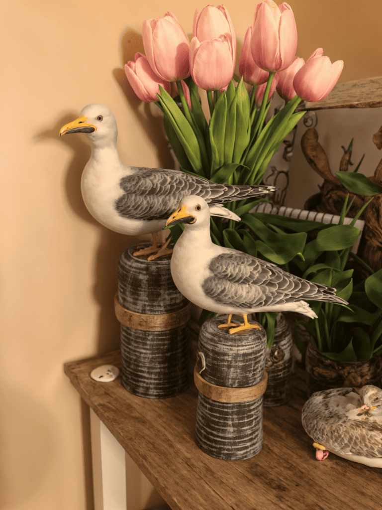 A shelf with two seagull statues sitting on wooden posts, and a vase of pink tulips in the background.
